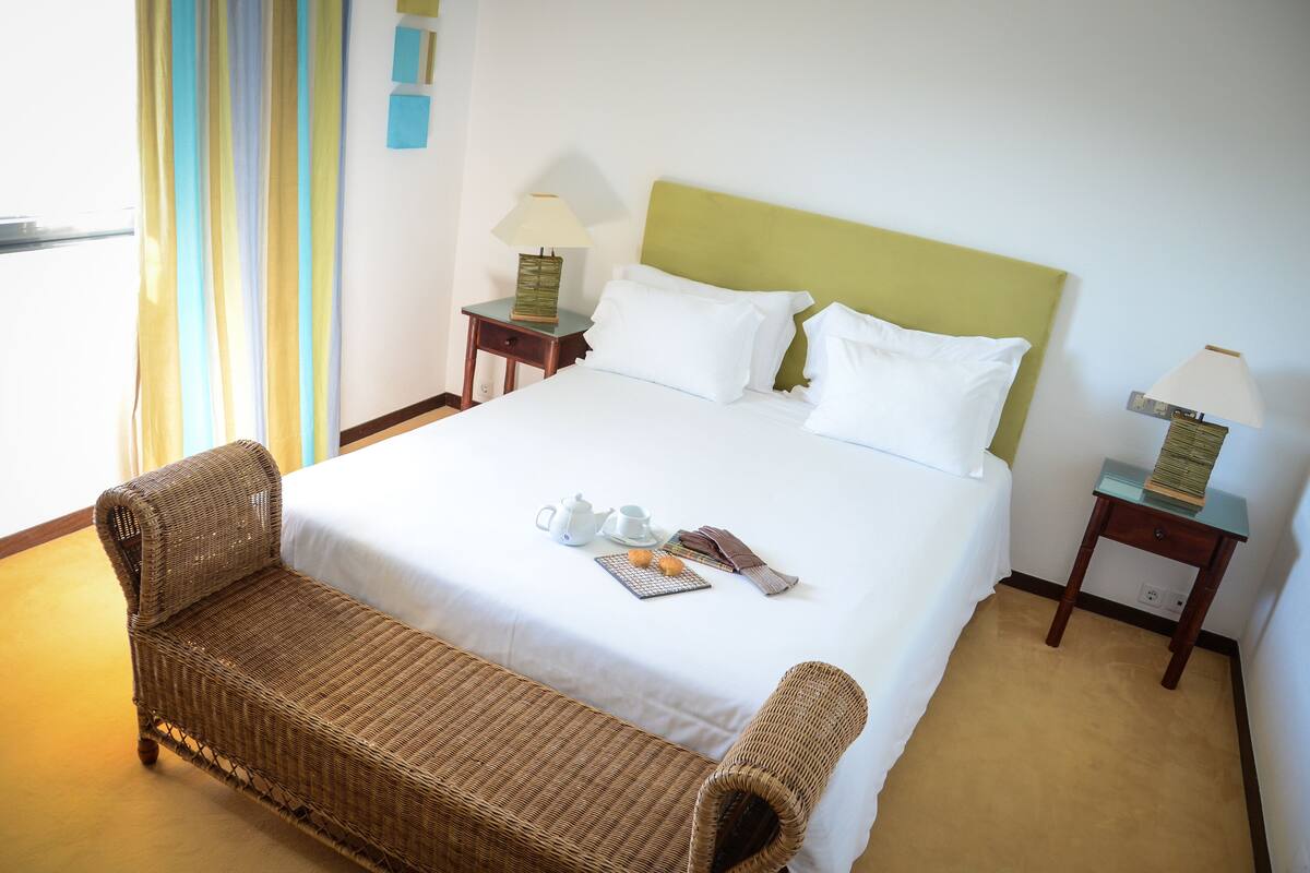 A neatly made bed with pillows, a bedside table with a lamp, a wicker chair, and a breakfast tray with a teapot and cups.