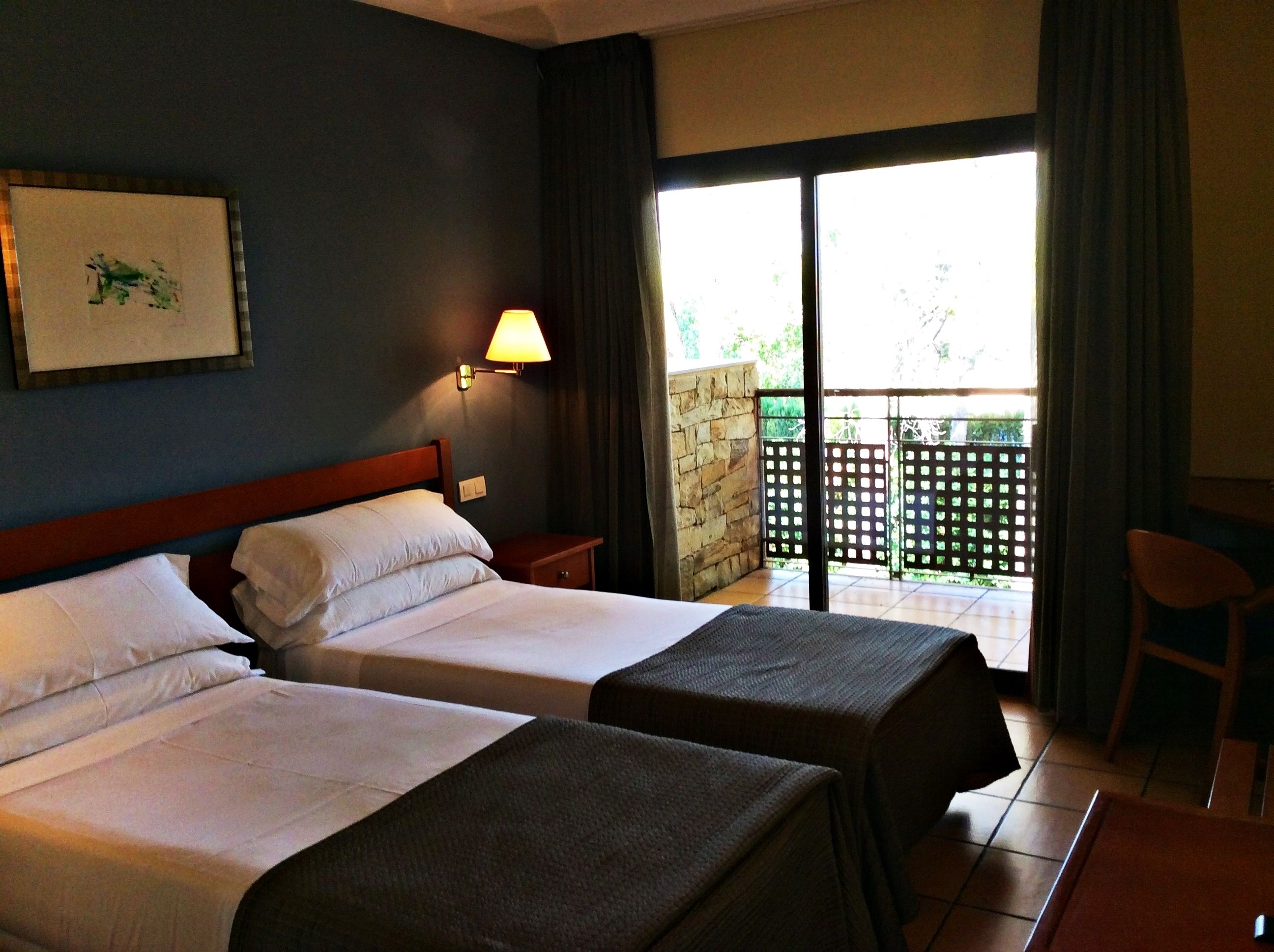 Habitación de hotel con dos camas, una mesita de noche, una lámpara y vistas al balcón exterior.