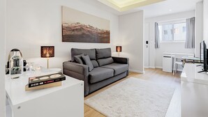 Una sala de estar moderna con un sofá gris, una mesa de centro y una estantería. Hay un cuadro en la pared y un escritorio con una silla.