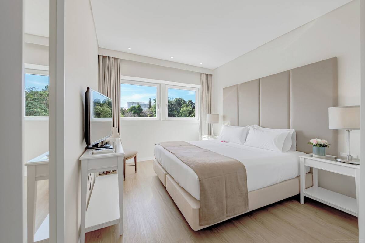Habitación de hotel moderna con una cama grande, un escritorio con computadora, televisión y ventana con vista a follaje.