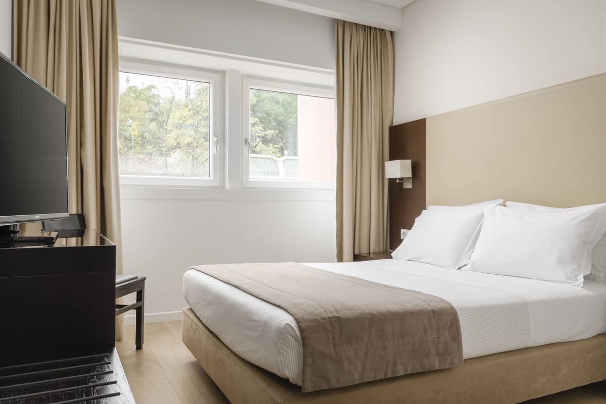 Una habitación de hotel con una cama, televisión y ventana con cortinas.