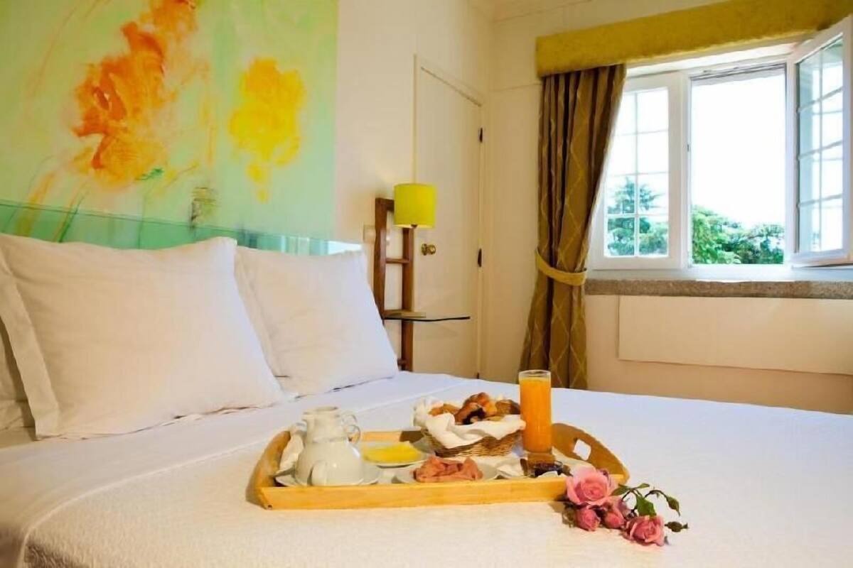 Habitación de hotel con cama, una bandeja con productos de desayuno, ventana con cortinas y un cuadro en la pared.