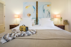 Een netjes opgemaakt bed met witte lakens, een gestreept plaid en een geweven tas ernaast. Twee nachtkastjes met lampen, een groot abstract schilderij aan de muur en een houten deur.