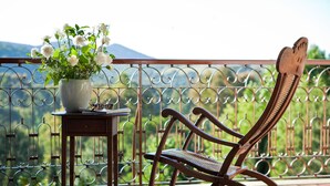Una silla de madera y una mesa con un florero, en un balcón con baranda metálica y vistas a zonas verdes y montañas.