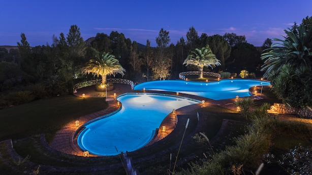 Una gran piscina serpenteante con un puente, rodeada de palmeras e iluminada con luces ambientales.