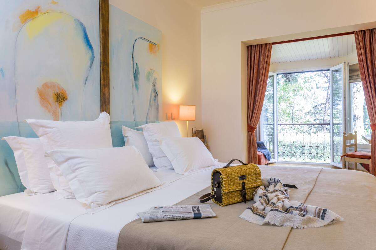 Habitación de hotel con una cama grande, ropa de cama blanca, un bolso tejido amarillo y un cuadro en la pared.