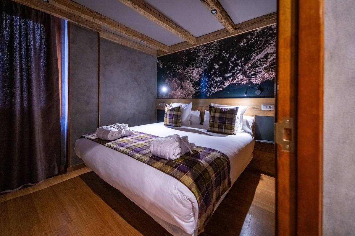 Quarto de hotel com uma cama grande, vigas de madeira no teto, um mural da noite estrelada na parede e piso de madeira.