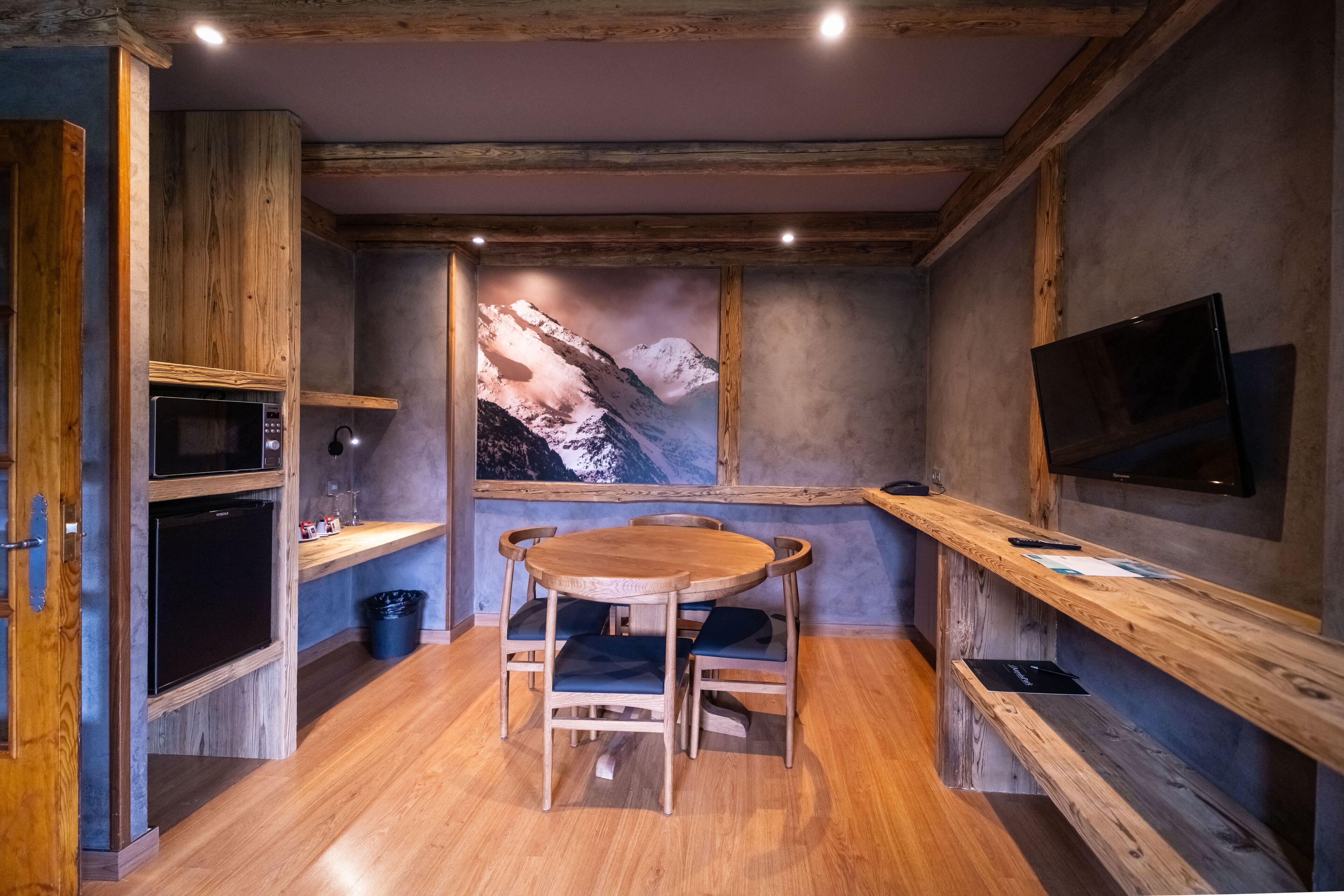 Una mesa de comedor de madera con sillas, un televisor en la pared y un cuadro con un paisaje de montaña.