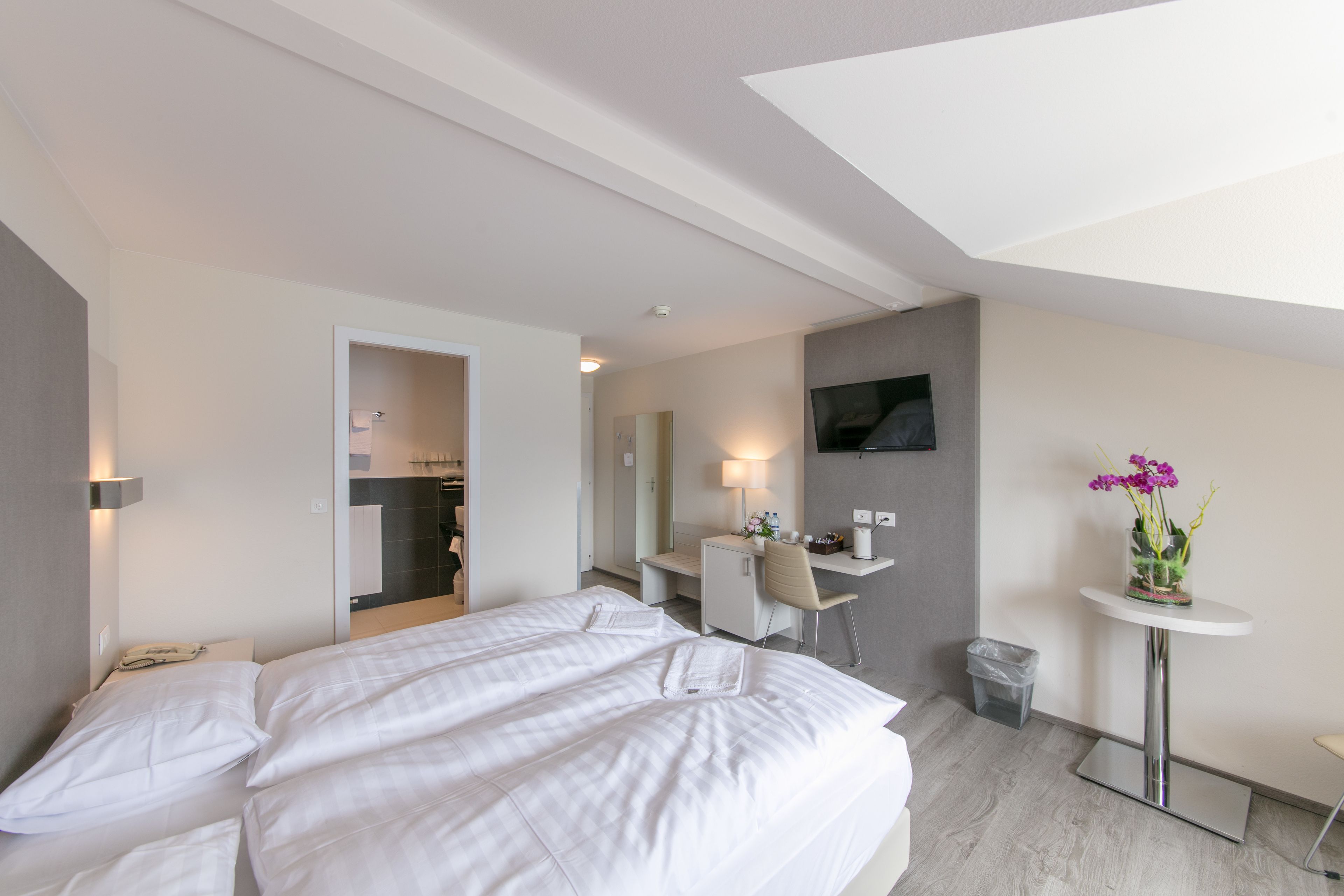 Une chambre d’hôtel avec un lit, un bureau, une chaise, une télévision et une petite table sur laquelle est posé un vase de fleurs.