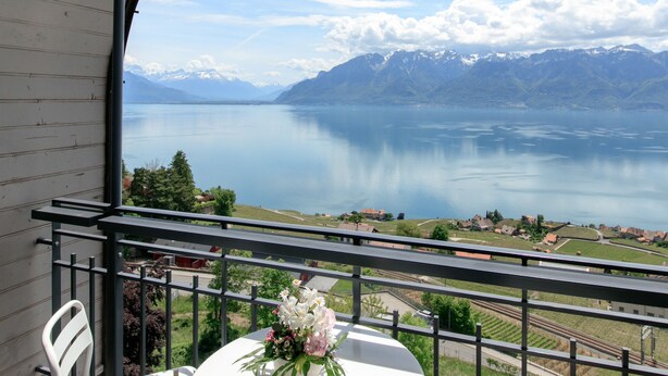 A balcony with a white table and chairs, overlooking a lake and mountains. - Hôtel Préalpina (Chexbres)