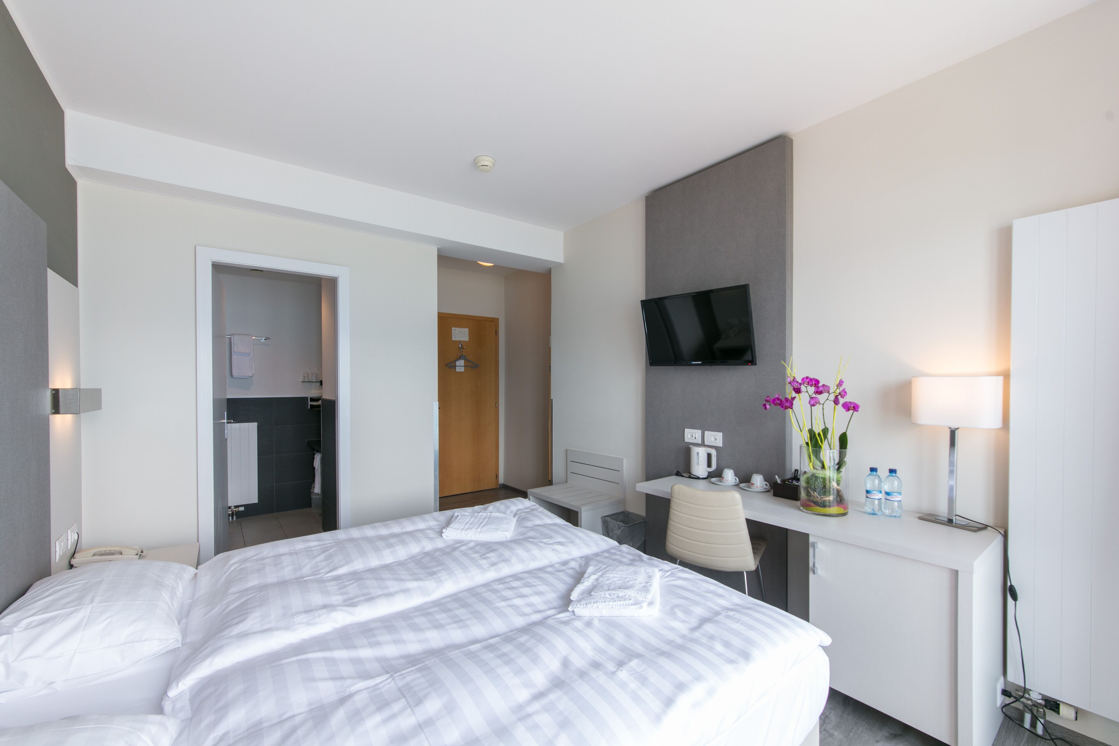 A hotel room with a large bed, a desk with a chair, a TV mounted on the wall, and a small table with a vase of flowers.