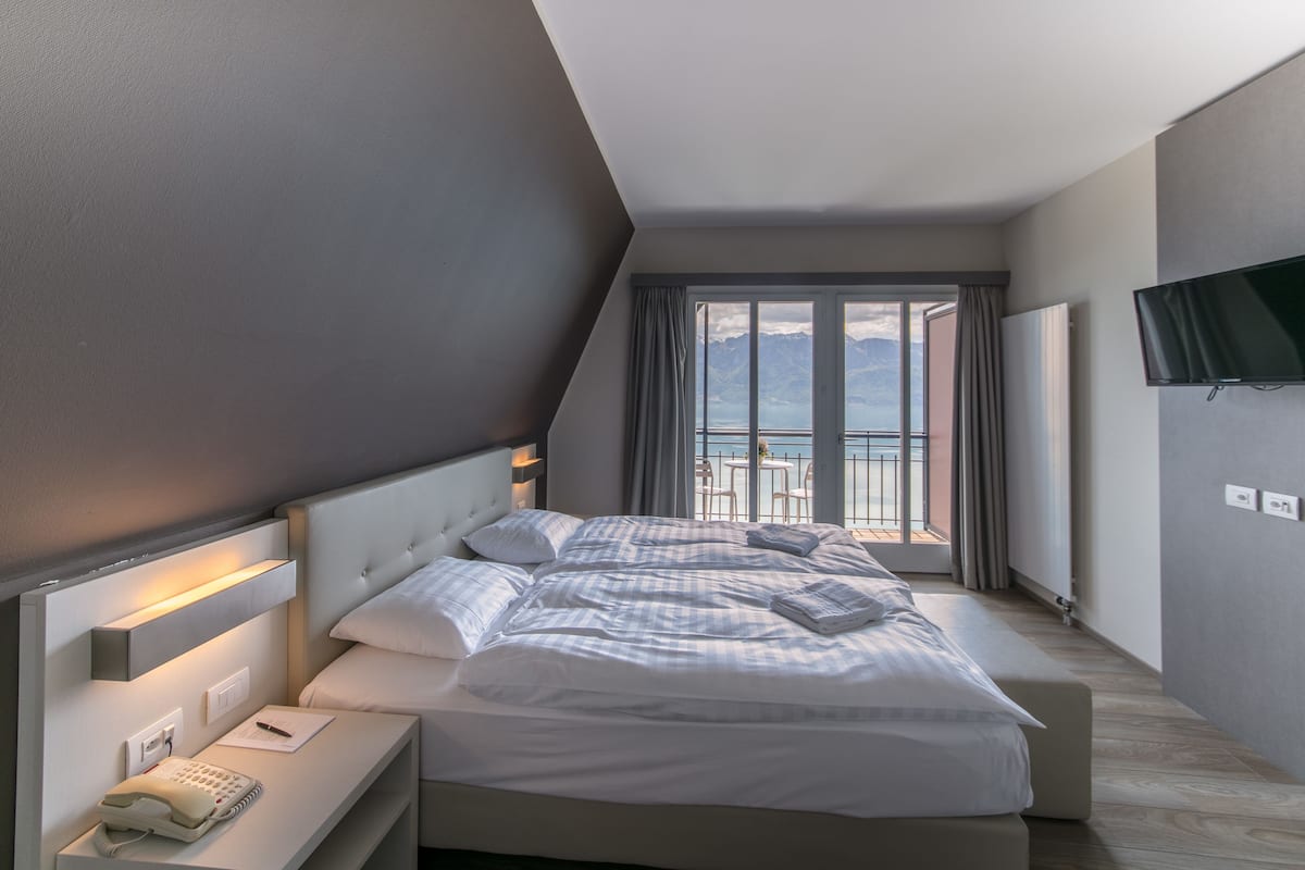 A modern hotel room with a large bed, a bedside telephone, and a view of the mountains through the window.