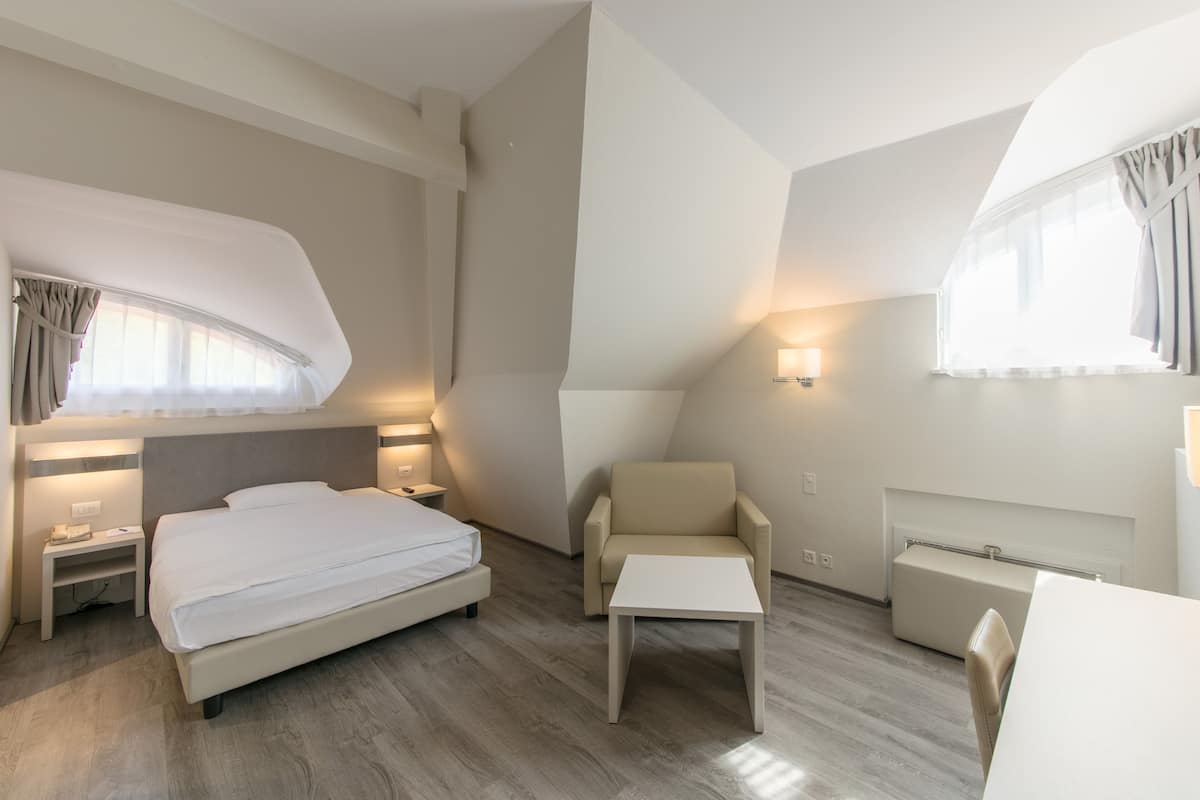 A modern hotel room with a large bed, a small desk, a chair, and a bedside table.