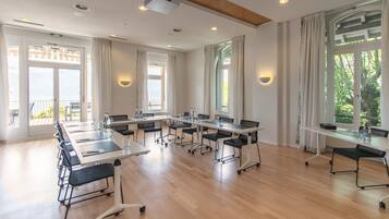 Un salón de conferencias con varias mesas y sillas, ventanas amplias con cortinas y un proyector en el techo.