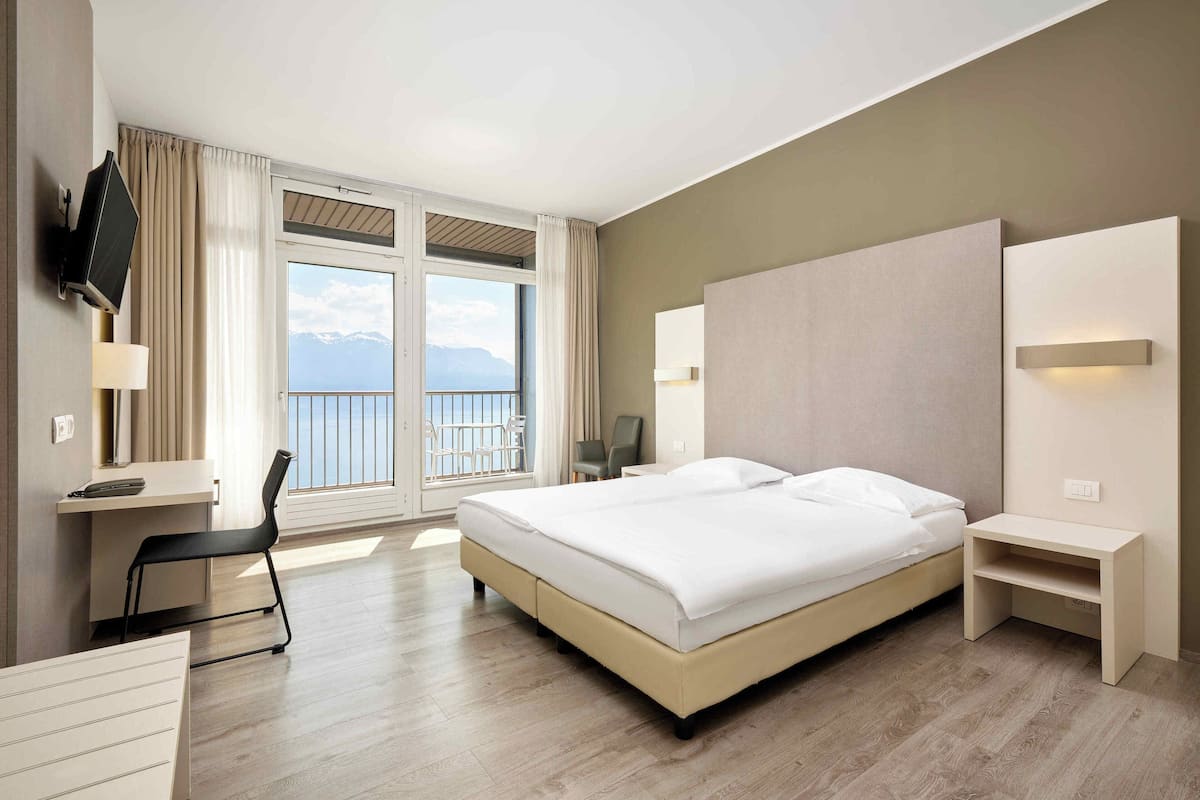 A hotel room with a large bed, a desk, a chair, a TV, and a view of the mountains.