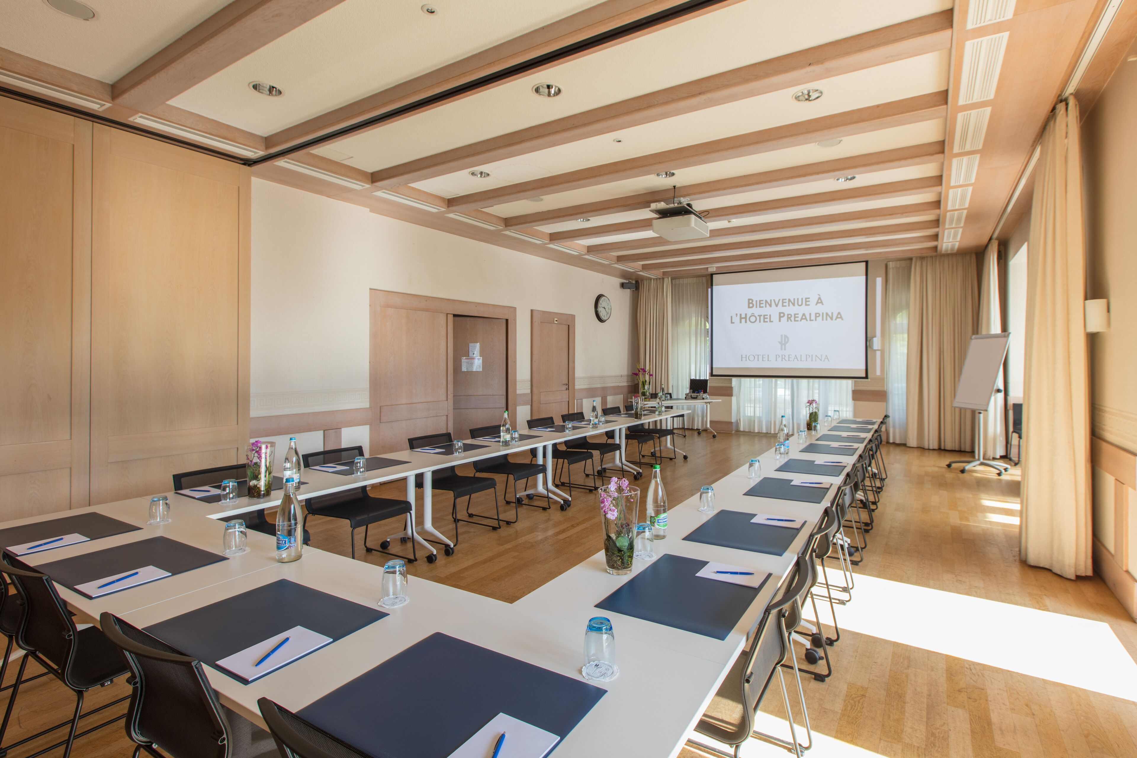 A conference room with long tables, chairs, a projector screen, and a clock on the wall.