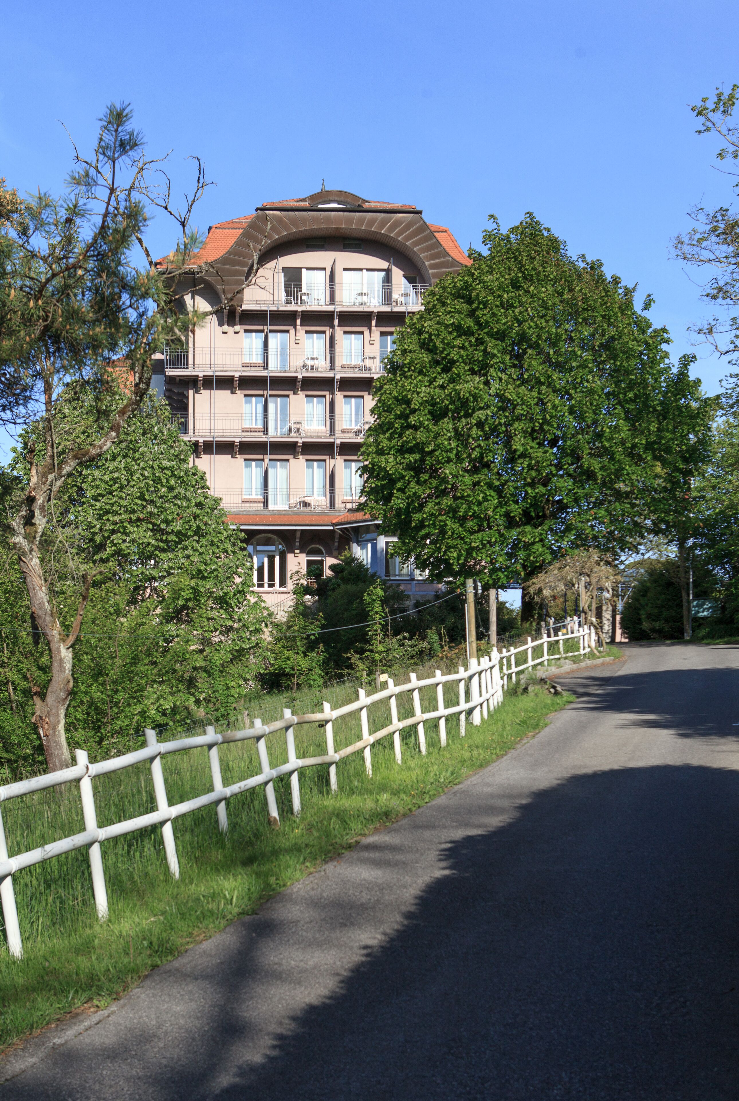 A multi-story building with balconies, surrounded by trees and a white fence along a curved road.
