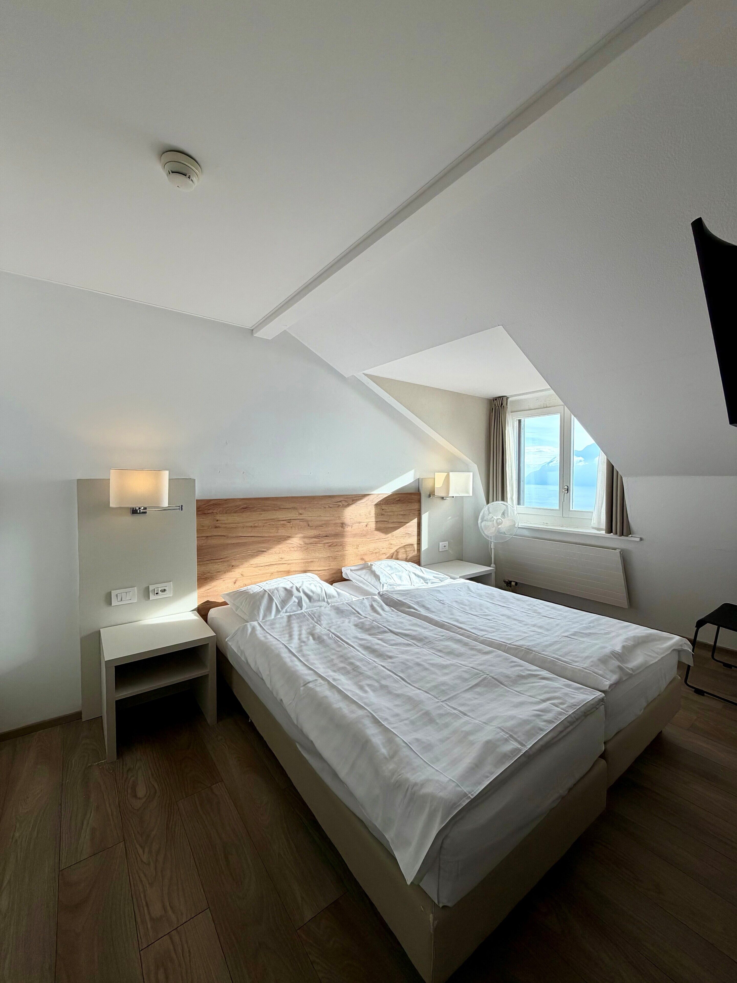 A modern bedroom with a large bed, a wooden headboard, bedside tables with lamps, a window with curtains, and a ceiling light.