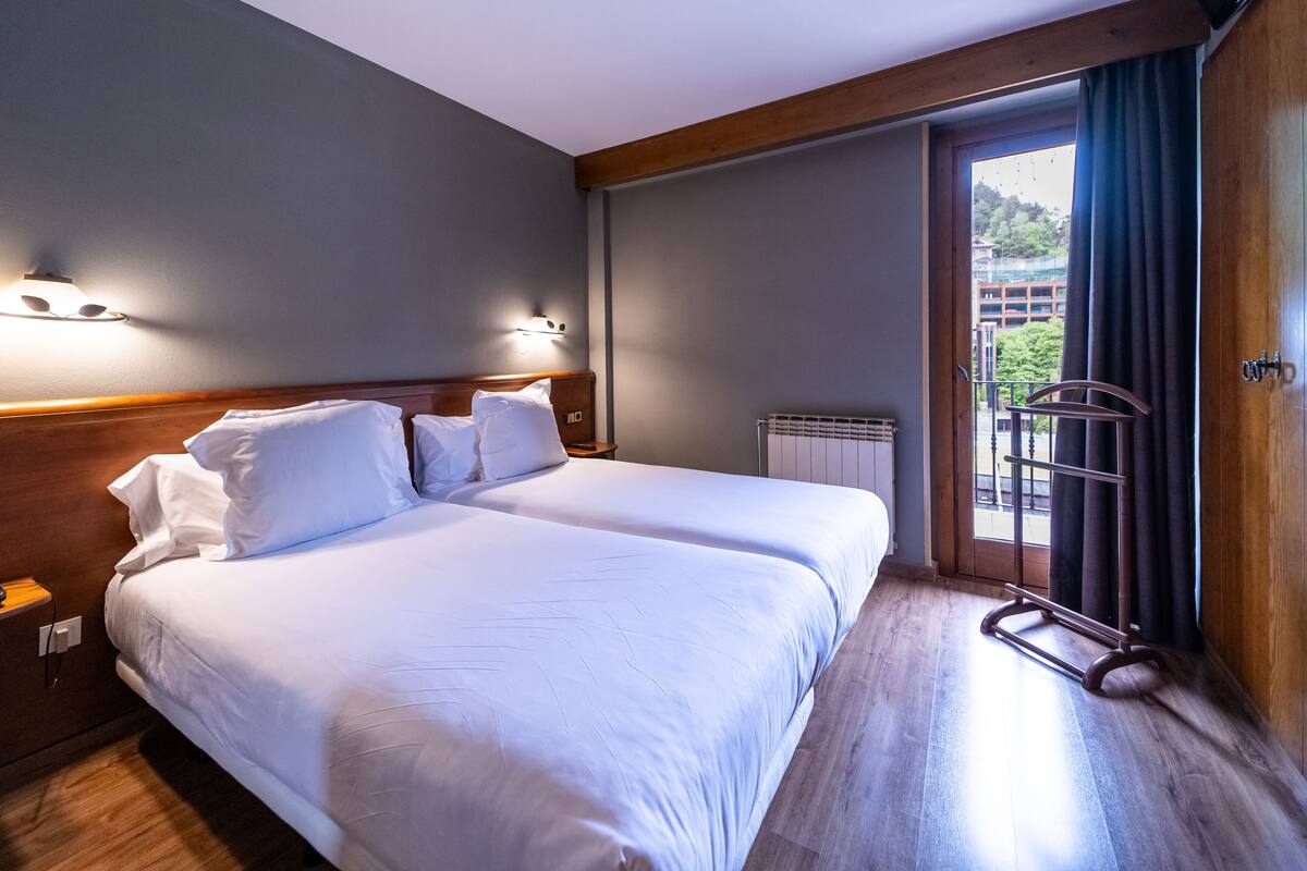 A hotel room with a double bed, a wooden headboard, a nightstand, a window with a view of buildings, and a balcony with a railing.