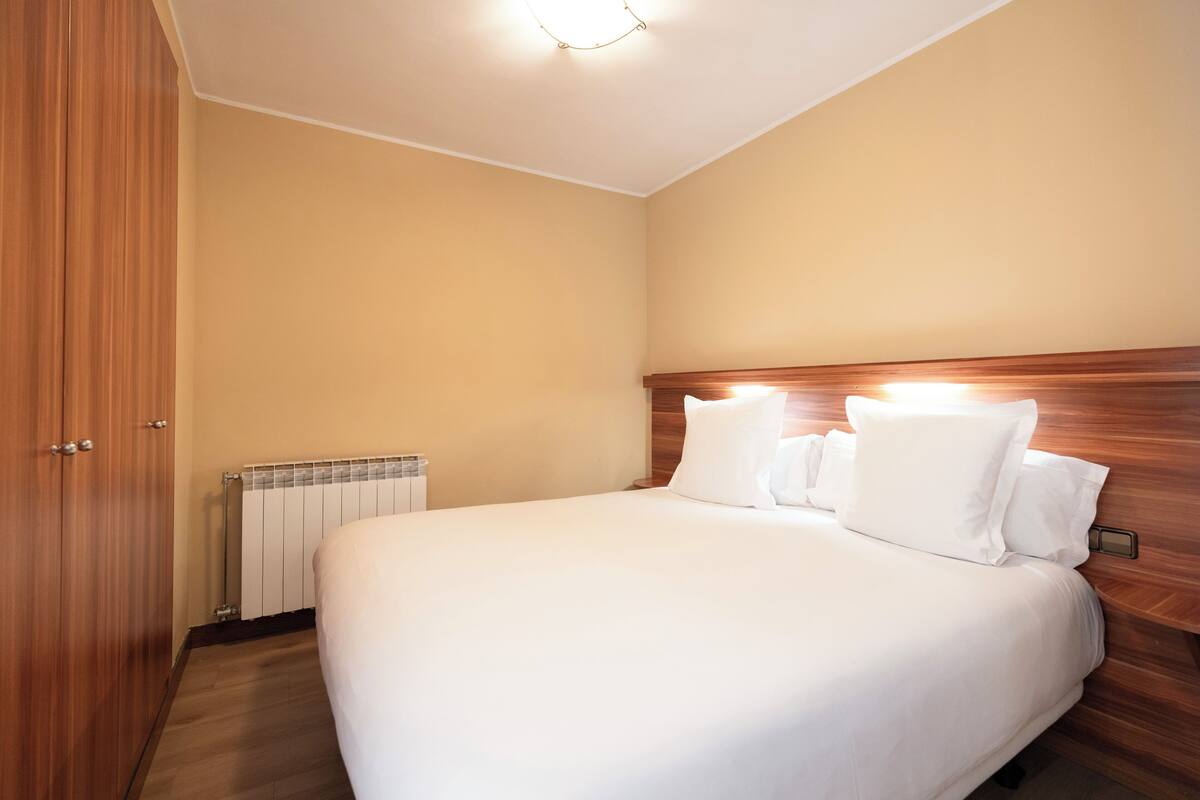 A hotel room with a wooden headboard, a large bed with white linens, a wooden wardrobe, and a radiator on the wall.