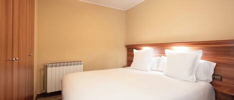 A hotel room with a wooden headboard, a large bed with white linens, a wooden wardrobe, and a radiator on the wall.
