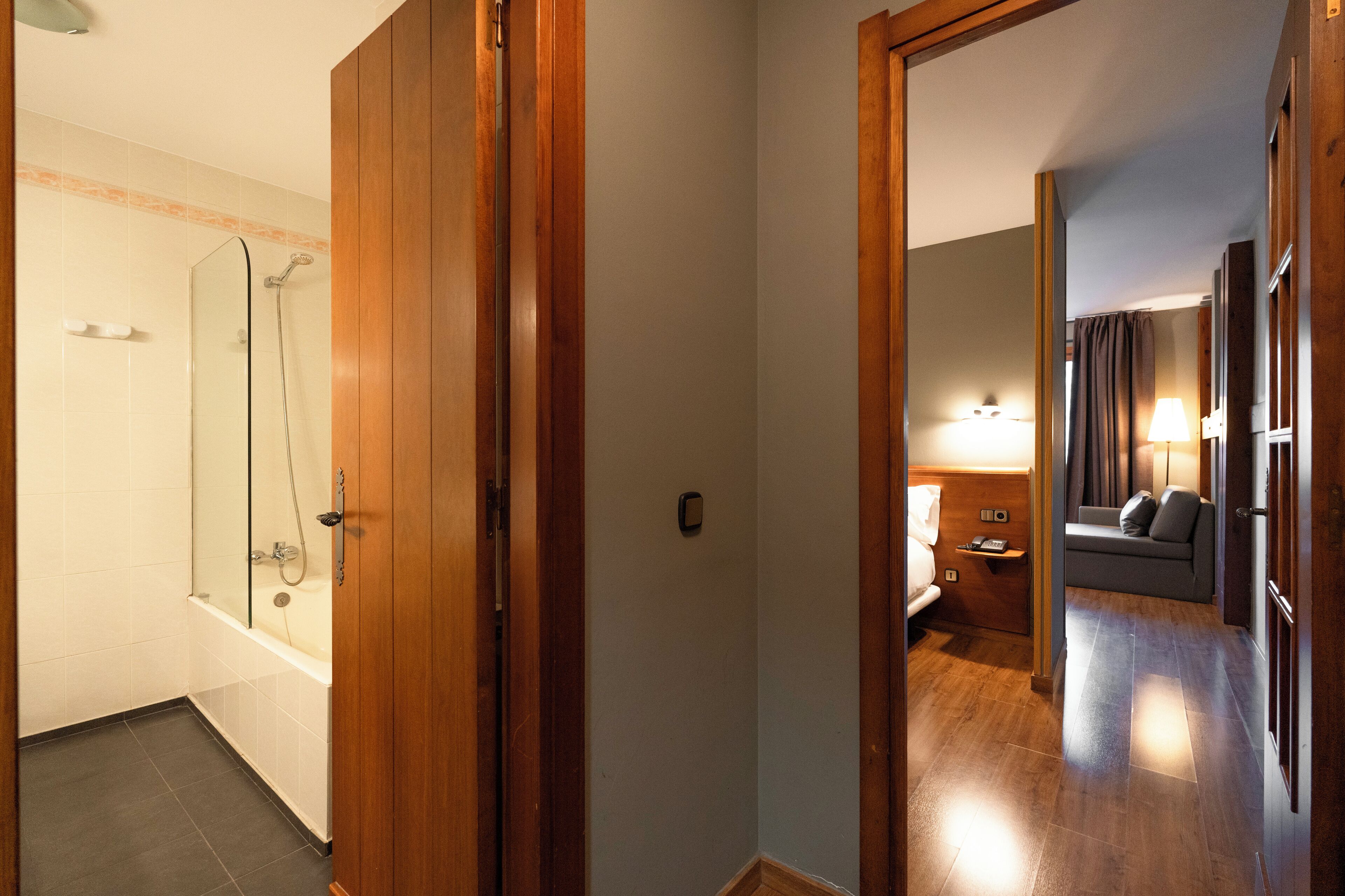 A hotel hallway with a bathroom on the left, a sofa and lamp in the center, and a wooden door on the right.