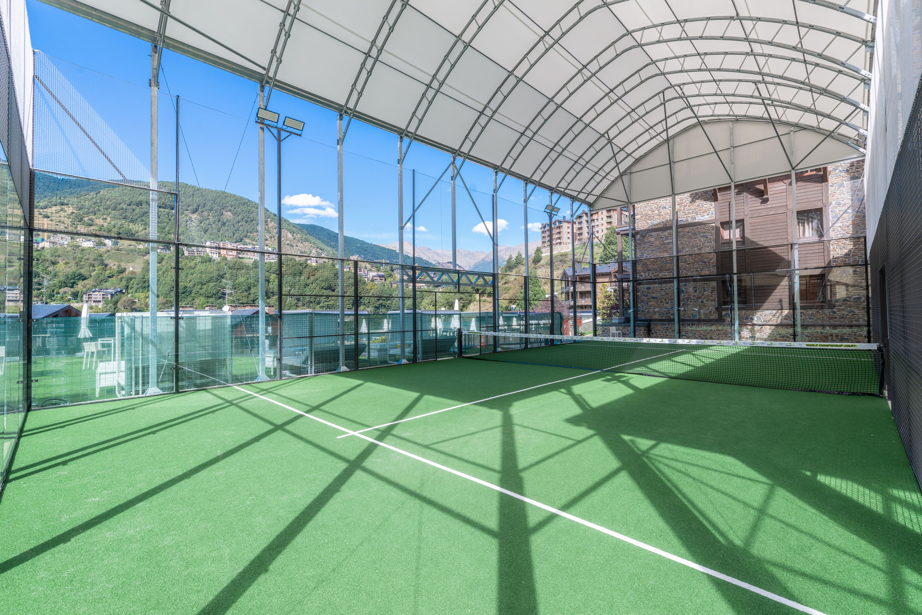An indoor tennis court with a glass enclosure, a green surface, and a view of a mountainous landscape.