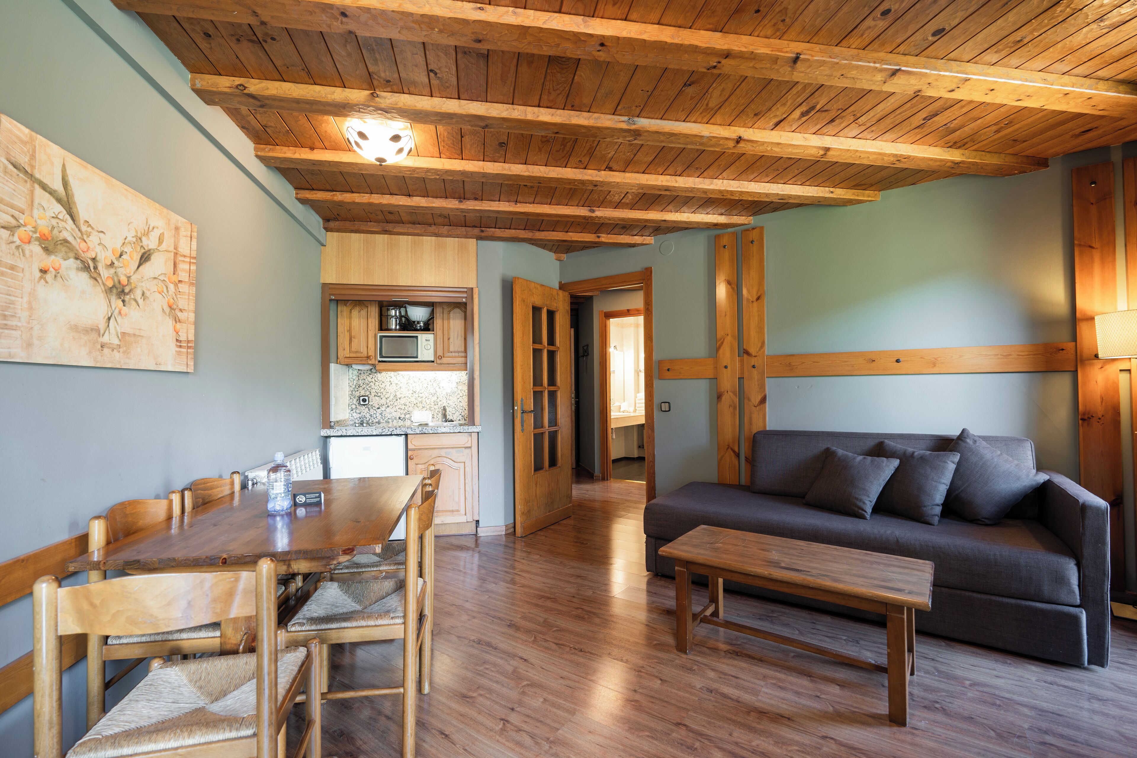 A wooden dining table with chairs, a sofa, and a kitchen area with cabinets and a microwave.
