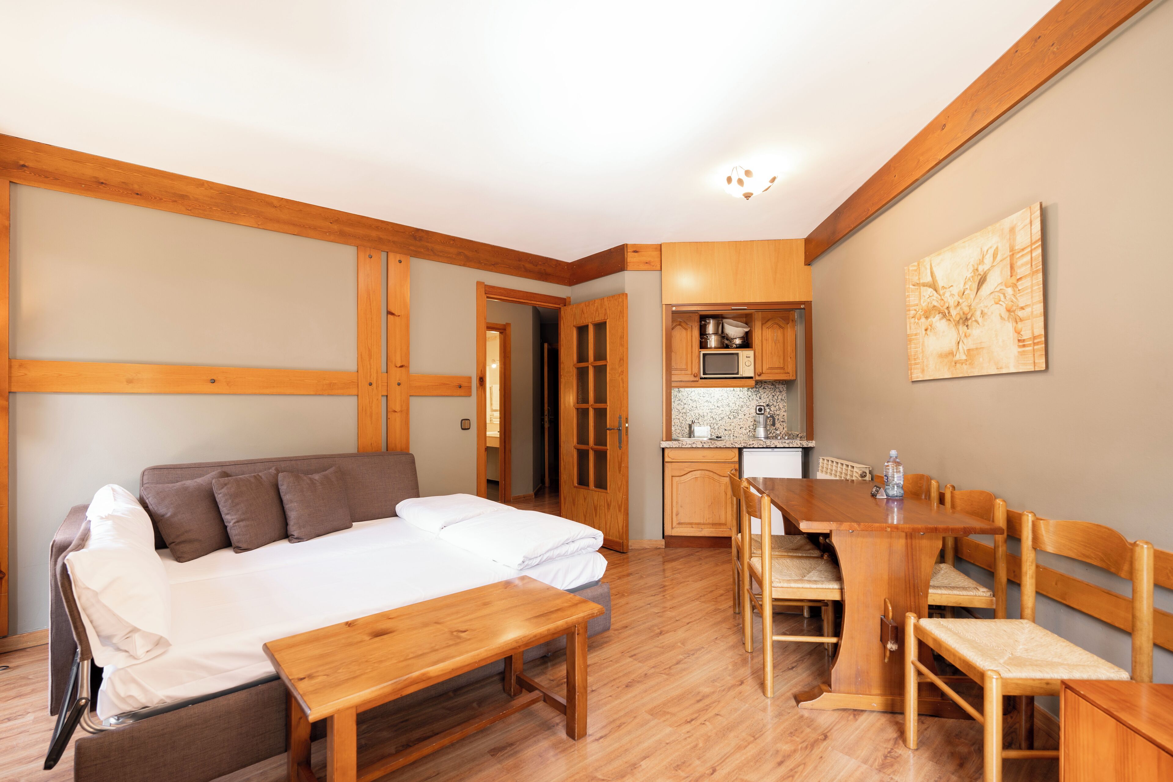 Habitación compacta con cama, banco de madera, mesa de comedor con sillas, zona de cocina y puerta que da a otra habitación.