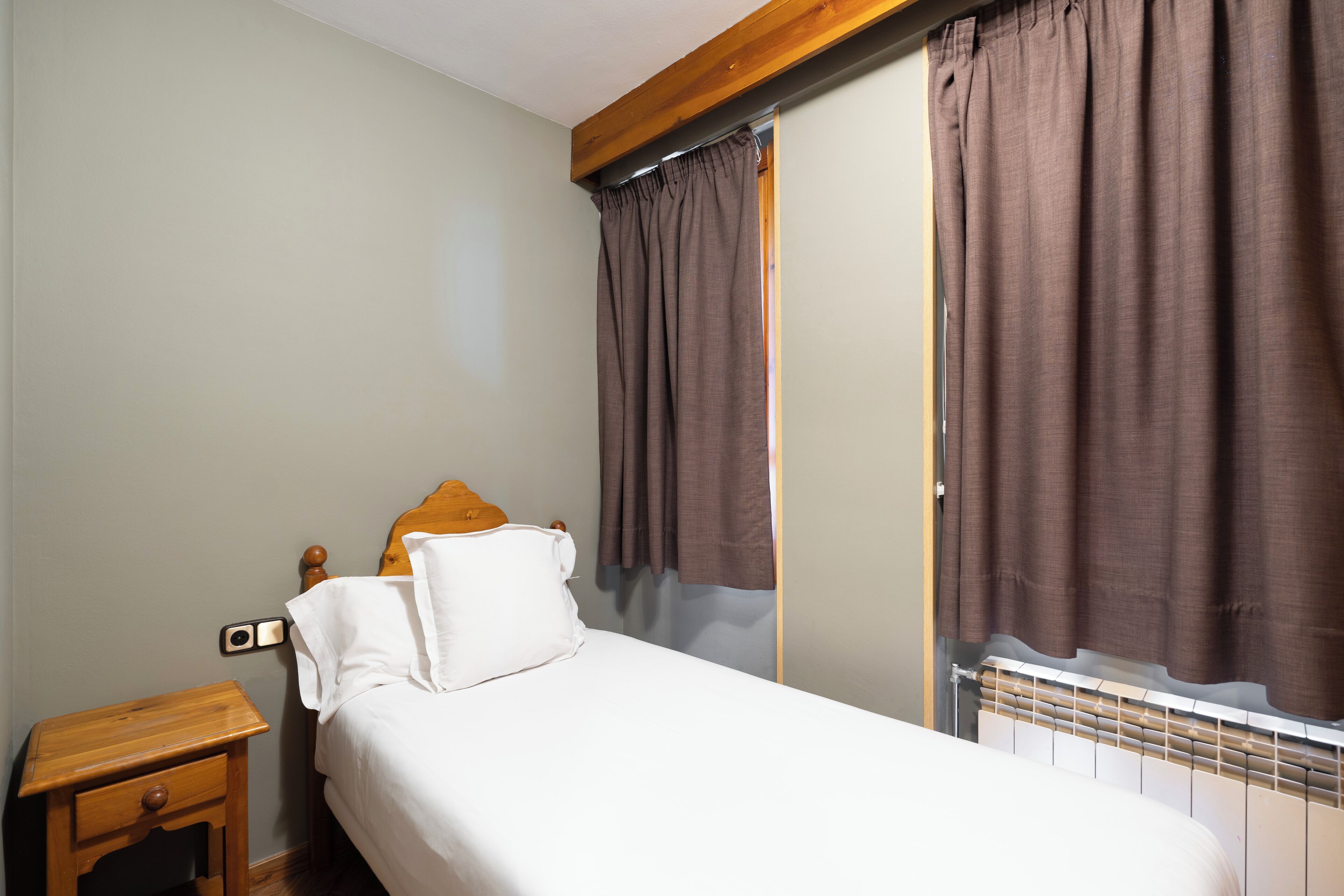 A bedroom with a bed, a wooden nightstand, a radiator, and brown curtains.
