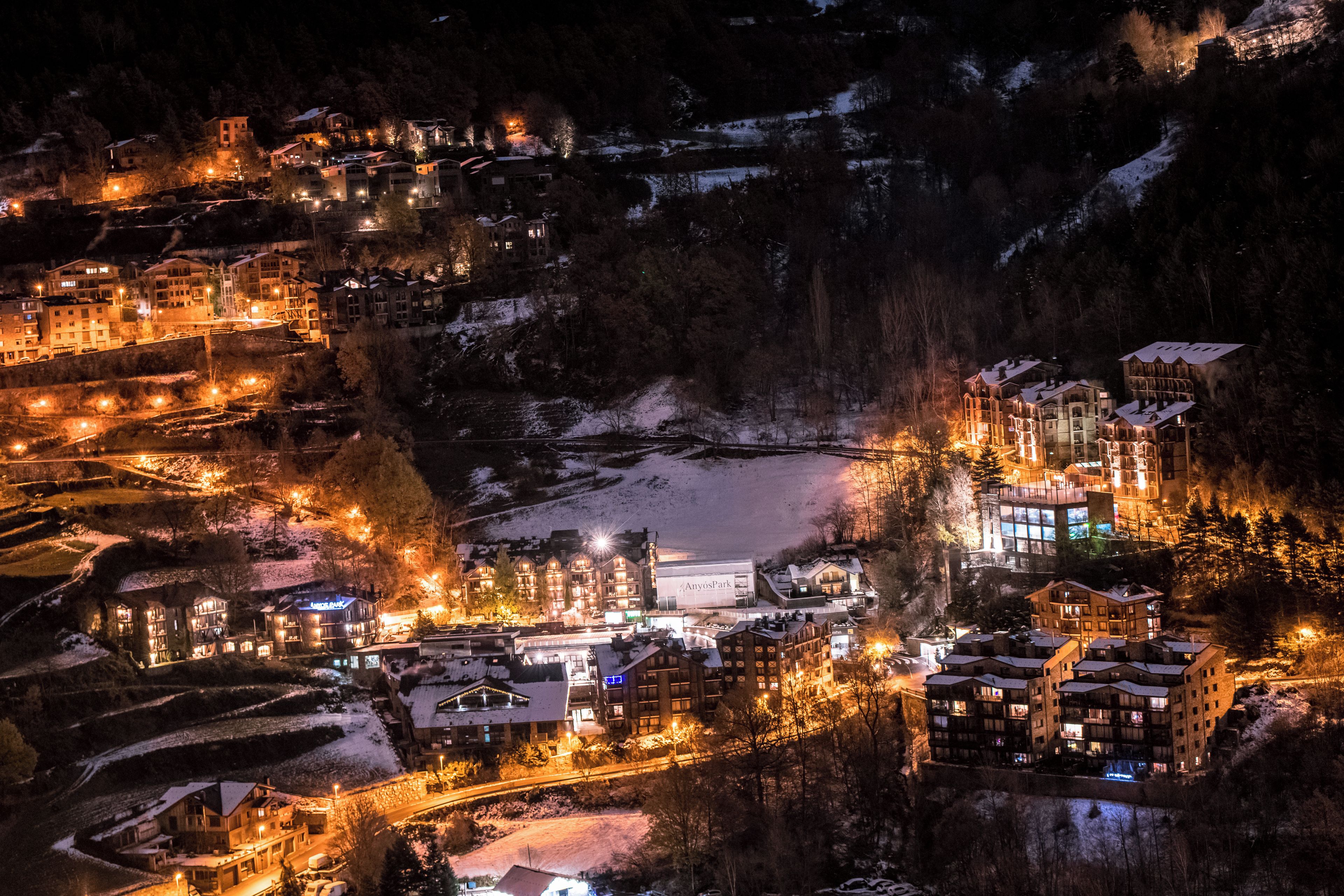 Un pueblo cubierto de nieve con edificios iluminados, un río y montañas al fondo.