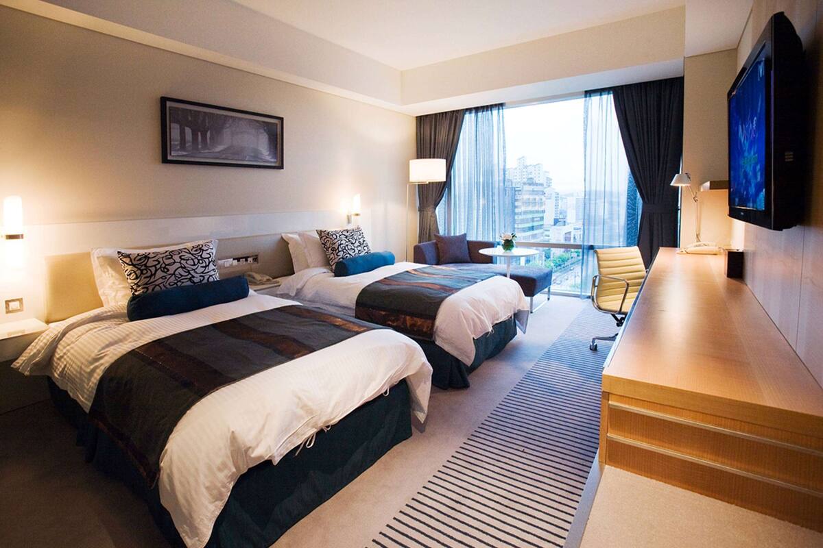 Habitación de hotel con dos camas, televisión, escritorio y vistas a la ciudad.