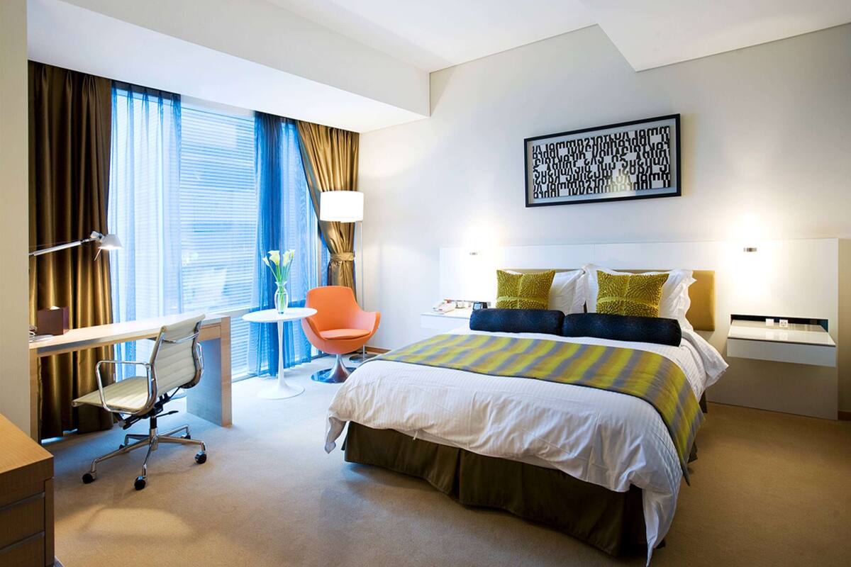 Habitación de hotel con cama, escritorio, silla y un ventanal con cortinas.