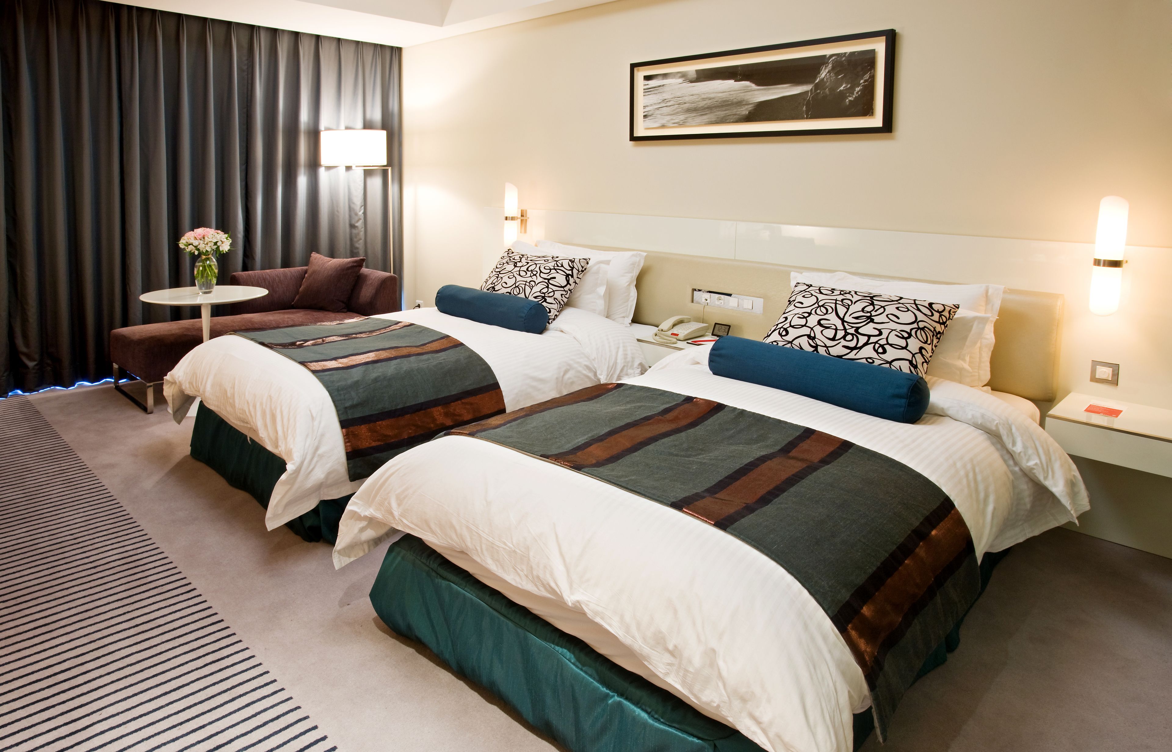 Habitación de hotel con dos camas, un sofá, una mesita con un florero y un cuadro en la pared.