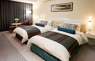 Habitación de hotel con dos camas, un sofá, una mesita con un florero y un cuadro en la pared.
