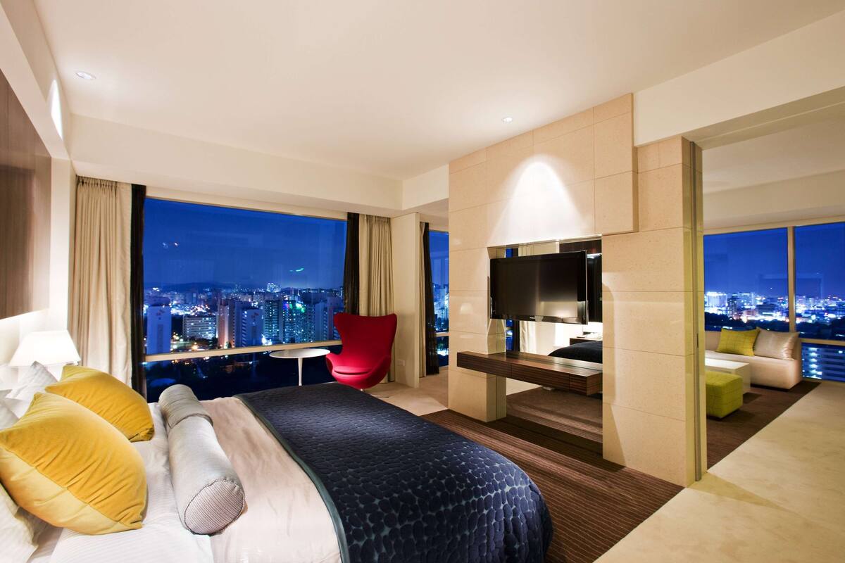 Una habitación de hotel moderna con una cama grande, una silla roja, un televisor y vistas a la ciudad.