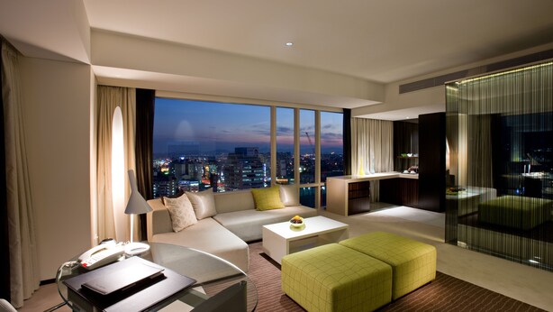 A modern hotel room with a city view, a sofa, a coffee table, a glass desk, and a green ottoman. - Ramada Plaza Suwon (Suwon)