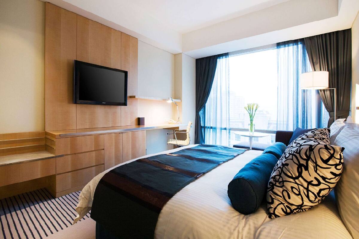 Habitación de hotel con cama, televisión montada en la pared, escritorio, lámpara y ventana con cortinas.