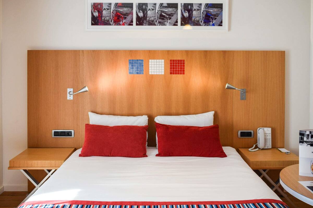 Habitación de hotel con cabecera de madera, dos almohadas rojas, una colcha a rayas, una mesita de noche con un teléfono y una pequeña mesa con una revista.
