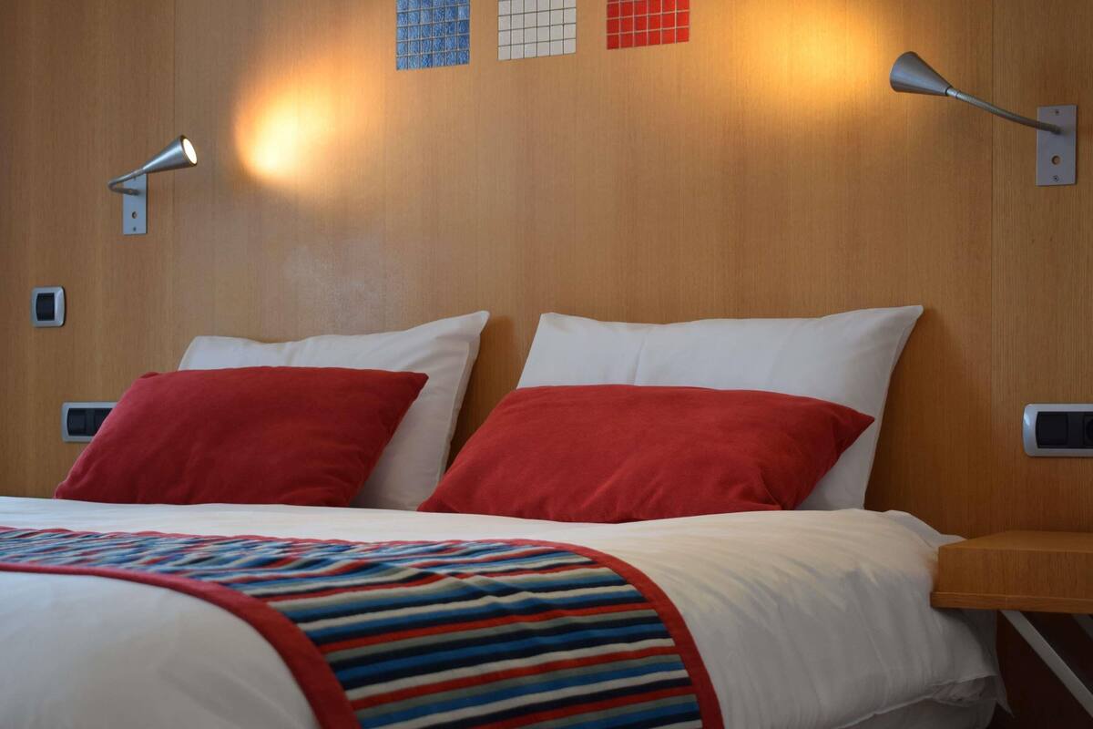 Habitación de hotel con cabecera de madera, dos almohadas rojas, una colcha a rayas y dos lámparas fijadas a la pared.