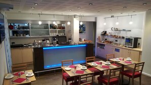 Un comedor moderno con mesas dispuestas para cuatro, una barra central con iluminación azul y una cocina al fondo.