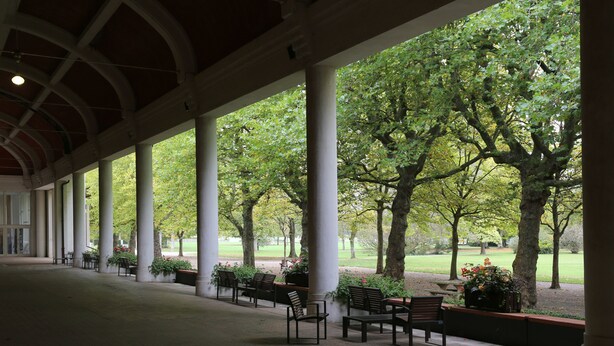 Un paseo techado con bancos y columnas, que conduce a un parque con árboles y césped.