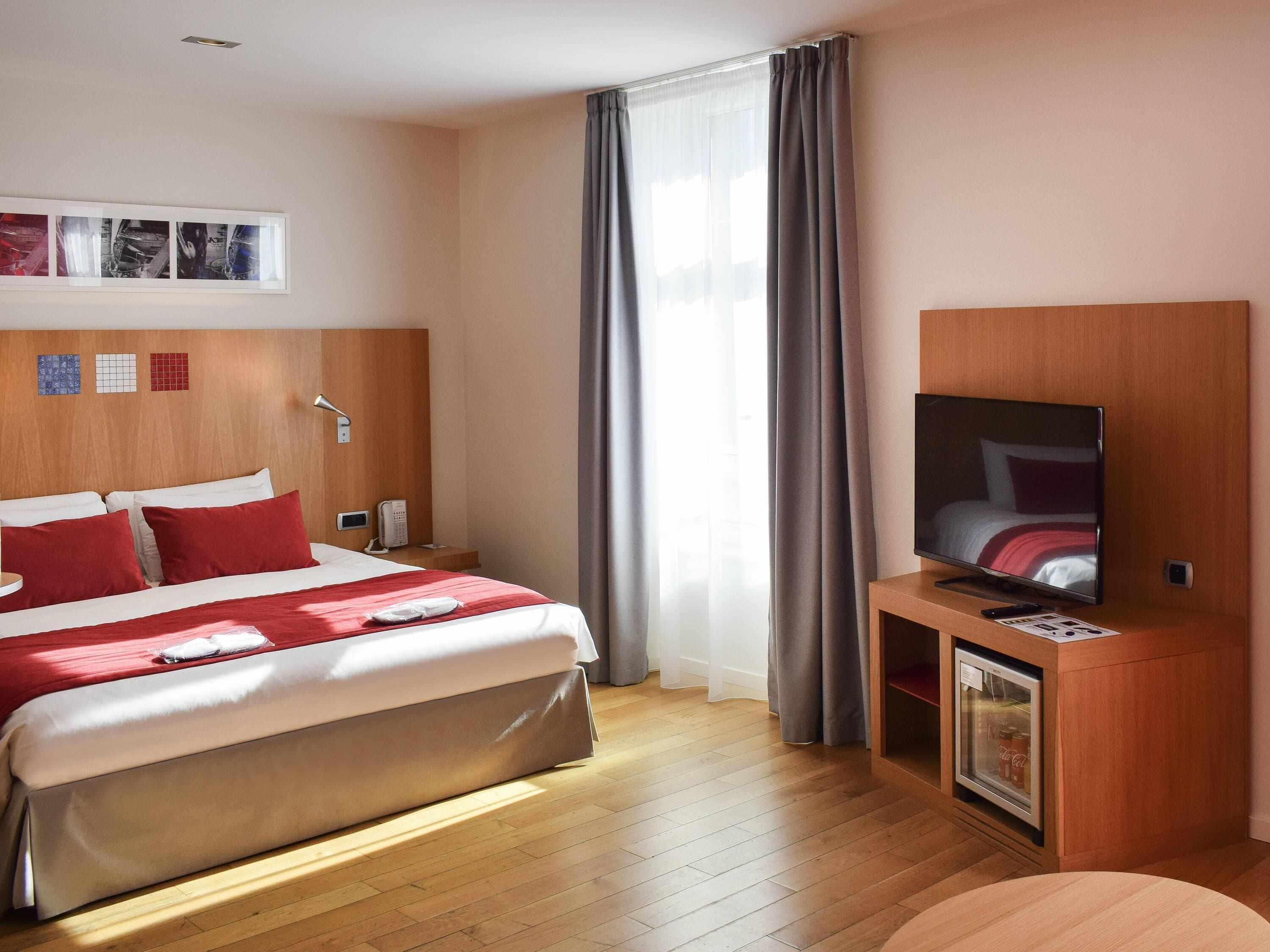 Een hotelkamer met een bed, een televisie, een minibar en een raam met gordijnen.