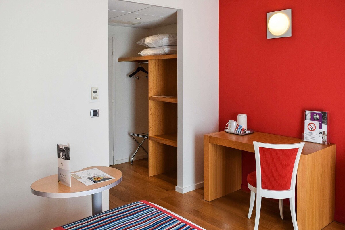 Une pièce avec un bureau en bois, un mur d’accent rouge, une chaise blanche, une petite table avec un magazine et un porte-manteau suspendu.