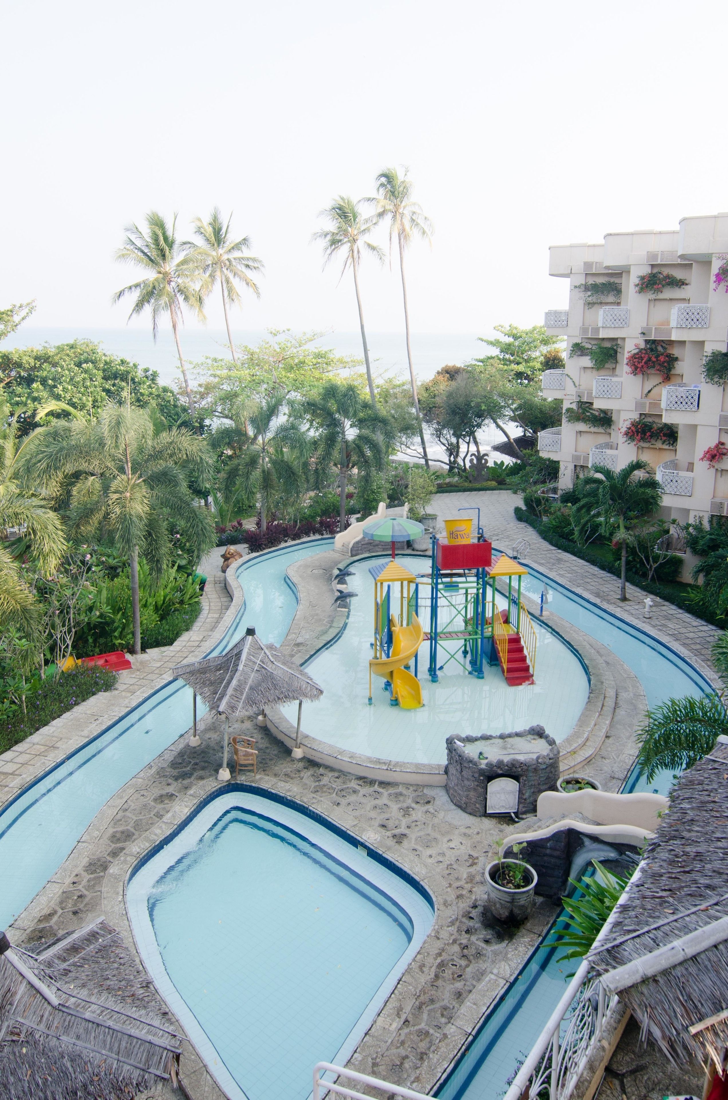 A pool area with multiple swimming pools, a children's play structure, and surrounding tropical vegetation.