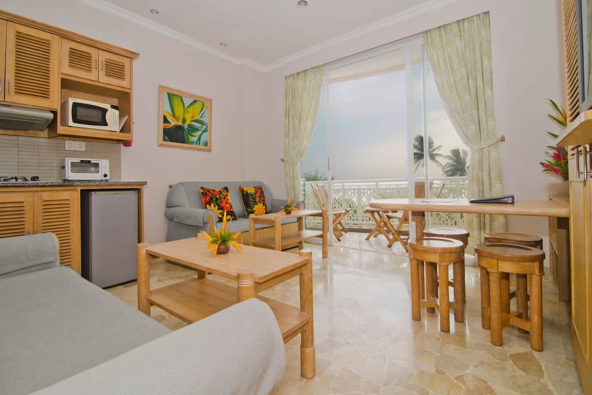 A modern living room with a sofa, a dining table with wooden chairs, a kitchen area with a microwave and a refrigerator, and a balcony with a view of palm trees.