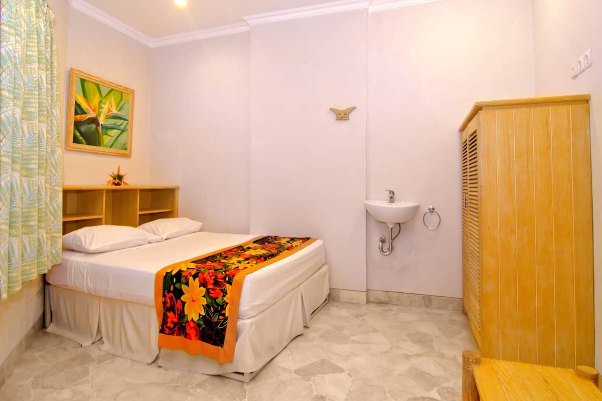 A bedroom with a bed, a bedside table, a nightstand, a window with curtains, a wall-mounted sink, and a wooden wardrobe.