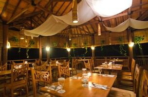Un restaurante con mesas y sillas de madera, cortinas blancas, plantas colgantes e iluminación colgante.