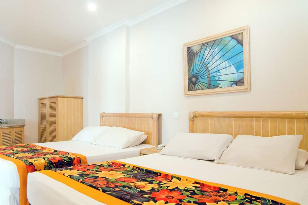 A hotel room with two beds, wooden headboards, a single bed with a colorful floral bedspread, a wooden wardrobe, and a wall-mounted abstract painting.