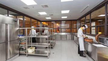 Une cuisine professionnelle équipée d’appareils en acier inoxydable, un chef cuisinier en train de préparer des plats et une grande fenêtre laissant entrer la lumière naturelle.